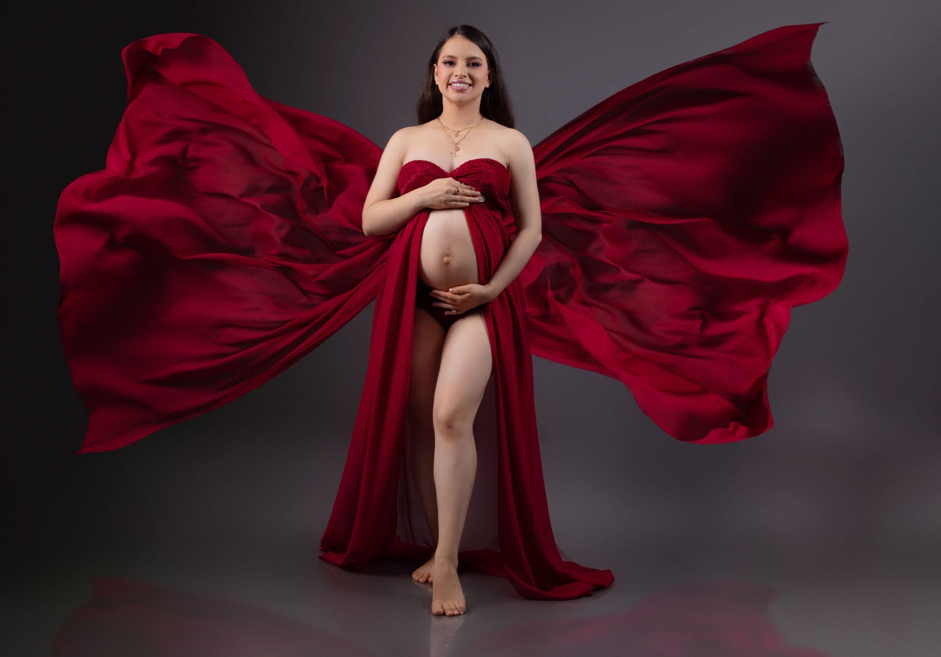 A pregnant woman in a long flowing dress, photographed against a plain backdrop.